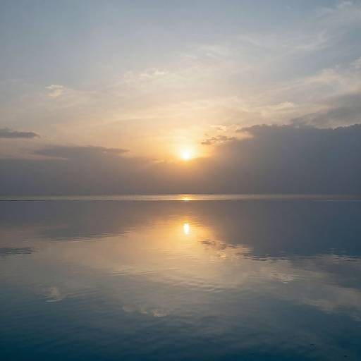 Photograph of a serene sunset over a calm, reflective ocean, with the sun partially obscured by clouds, casting a golden glow.