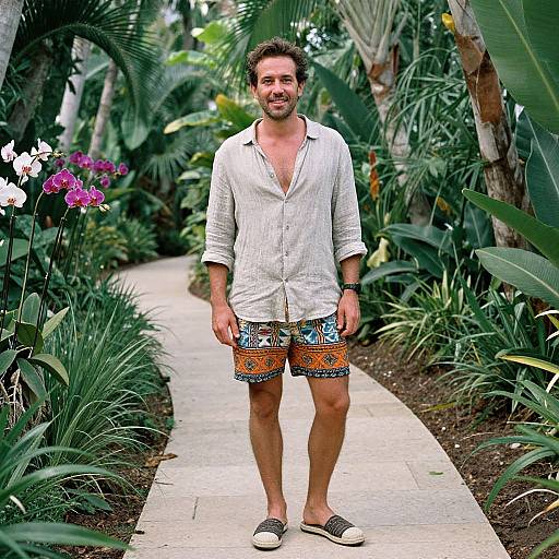 Photograph of a smiling, bearded man in a white button-up shirt and colorful shorts, standing on a garden path surrounded by lush tropical plants and