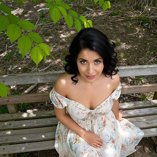 Photograph of a smiling, dark-haired woman with curly hair, wearing an off-shoulder floral dress, sitting on a wooden bench in a sun
