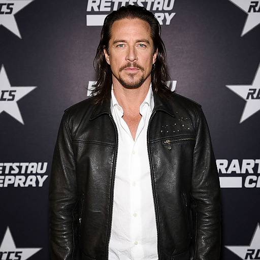 Photograph of a man with long dark hair, light beard, wearing a black leather jacket over a white shirt, standing against a black 
