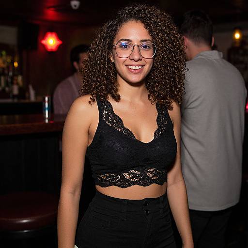 Photograph of a smiling woman with curly brown hair, wearing glasses, black lace crop top, and high-waisted black pants, in a dim
