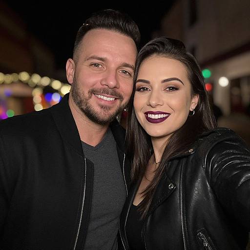Photograph of a smiling couple at night; man with short dark hair and beard in black jacket, woman with dark hair, black leather jacket, and