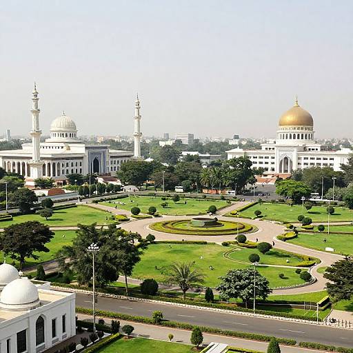 Photograph of a lush, green, well-maintained garden with winding paths, leading to grand white and gold-domed mosques with tall minare