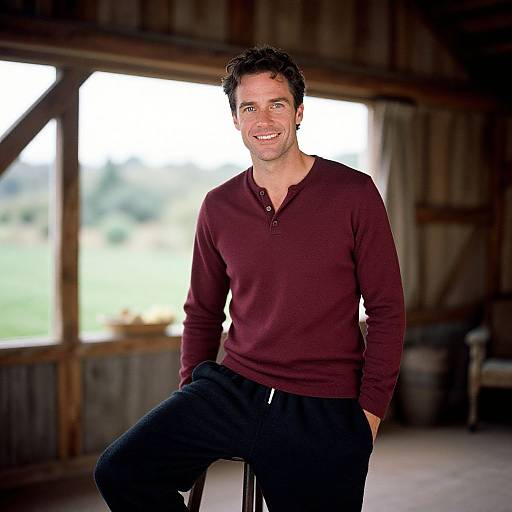 Photograph of a smiling, medium-built man with short dark hair, wearing a maroon long-sleeve henley and black pants, standing in