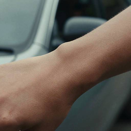Close-up photograph of a light-skinned, slightly veined forearm resting on the steering wheel of a car with a blurred, gray vehicle interior in the
