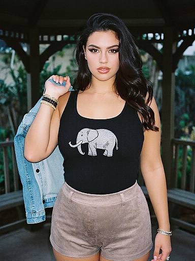 Photograph of a curvy woman with long black hair, wearing a black elephant tank top, beige shorts, holding a blue denim jacket, standing on