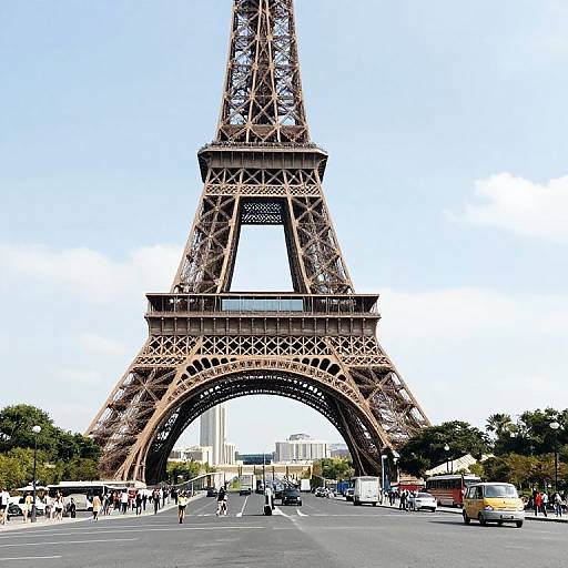 Photograph of the Eiffel Tower archway in Paris, with bustling street below, people walking, cars driving, and clear blue sky.