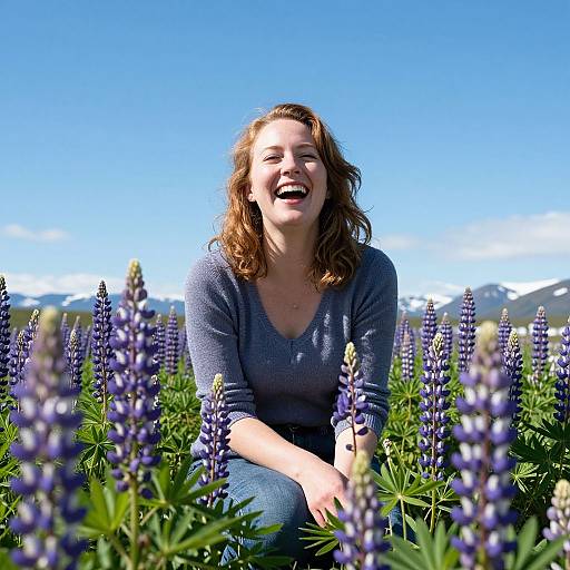 Photograph of a smiling, fair-skinned woman with wavy brown hair, wearing a blue sweater, kneeling in a vibrant lavender field under a clear