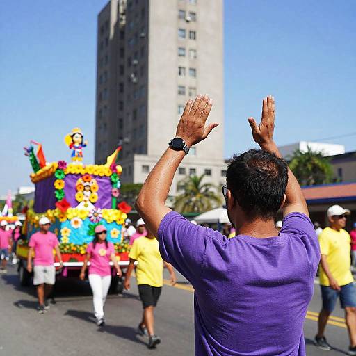 Photograph of a man in a purple shirt, hands raised, watching a colorful, float-filled parade with people in yellow and pink shirts. Tall building