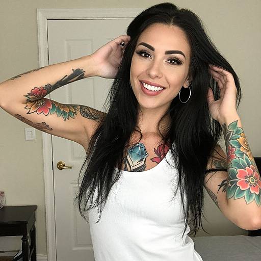 Photograph of a smiling woman with long black hair, tattoos on arms and chest, wearing a white tank top, standing in a beige room with a