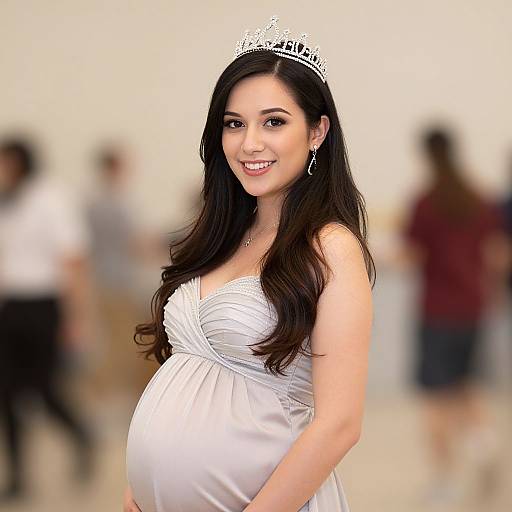Photograph of a smiling, pregnant woman with long, dark brown hair, wearing a white strapless dress and silver tiara, standing in a blurred
