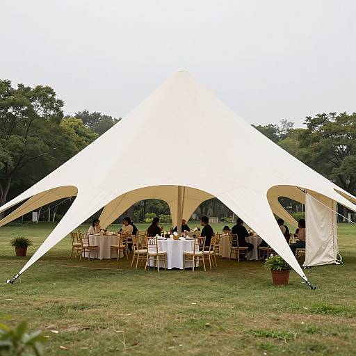 Photograph of an outdoor white tent with arched sides, hosting a formal dinner party on a grassy field surrounded by trees. Guests in black attire