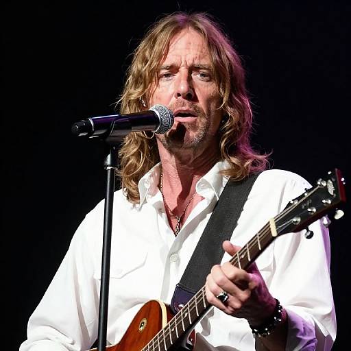 Photograph of an older man with long blonde hair, white shirt, and beard, singing into a microphone while playing an electric guitar on stage.