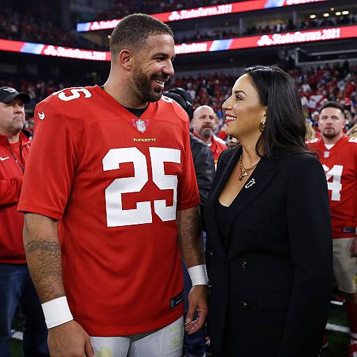 Photograph of a smiling African-American male football player in a red jersey number 25, standing beside a smiling Latina woman in a black blazer on