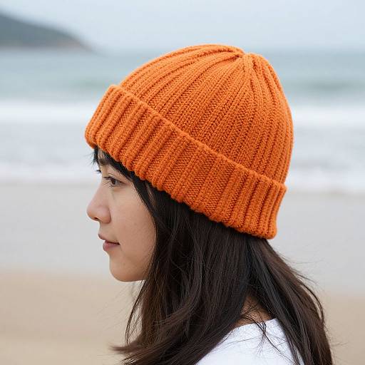Photograph of an Asian woman with long black hair wearing an orange knitted beanie, profile view, standing on a sandy beach with ocean and blurred