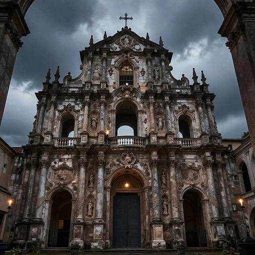 Photograph of a grand, ornate Baroque-style church facade under dramatic, cloudy skies. Detailed stone carvings, three arched openings,