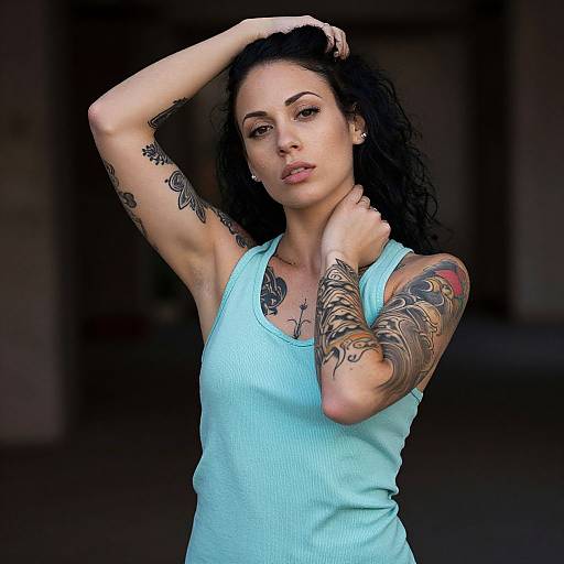 Photograph of a tattooed woman with curly black hair, wearing a light blue tank top, posing with one arm raised, against a dark, blurred