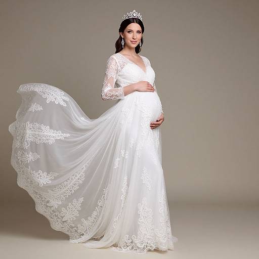 Photograph of a pregnant woman in a white lace wedding gown with long sleeves and a flowing train, wearing a silver tiara, standing against a plain