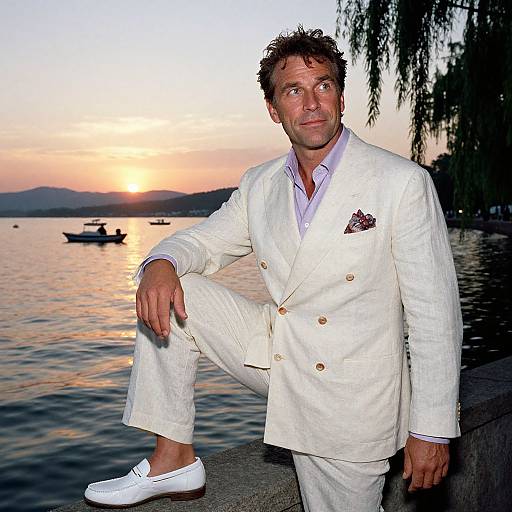 Photograph of a handsome man with curly brown hair in a white double-breasted suit, leaning on a lake's edge at sunset.