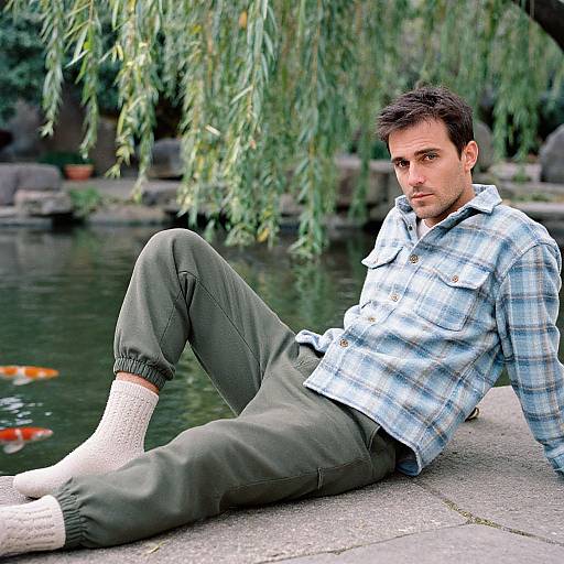 Photograph of a young man with short dark hair, wearing a blue plaid shirt and green pants, sitting by a pond with koi fish,