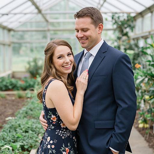 Photograph of a smiling couple in a greenhouse; woman in floral dress, man in navy suit, standing closely, touching lapels.