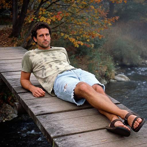 Photograph of a relaxed, short-haired man in a white t-shirt and frayed denim shorts, lounging on a wooden dock, wearing brown sandals