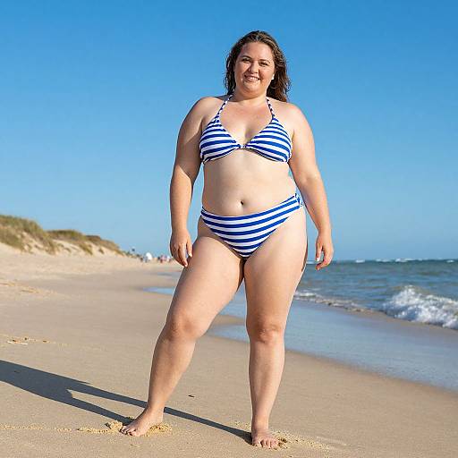 Photograph of a smiling, plus-sized woman with fair skin, dark hair, wearing a blue and white striped bikini, walking on a sunny, sandy