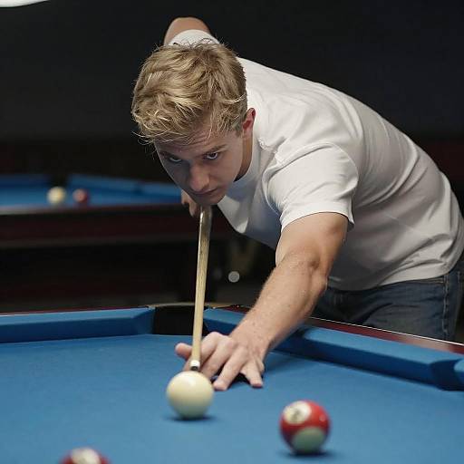 Photograph of a focused, blonde man in a white t-shirt, aiming a pool cue at white and red billiard balls on a blue-felt