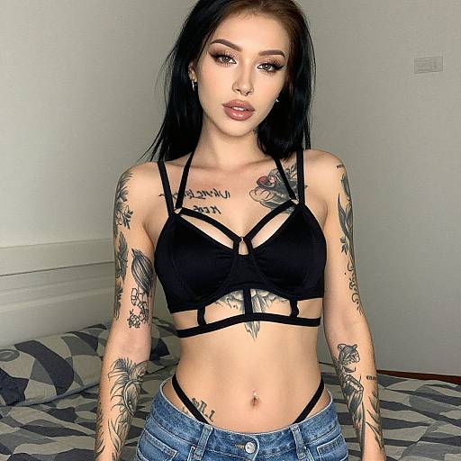 Photograph of a tattooed, pale-skinned woman with black hair, wearing a black strappy bra and blue jeans, standing in a bedroom with
