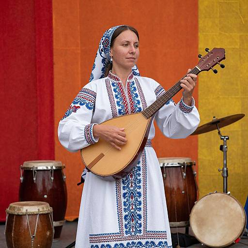 Photograph of a woman in traditional white embroidered dress with blue and red patterns, playing a banjo, standing in front of vibrant red and yellow curtains