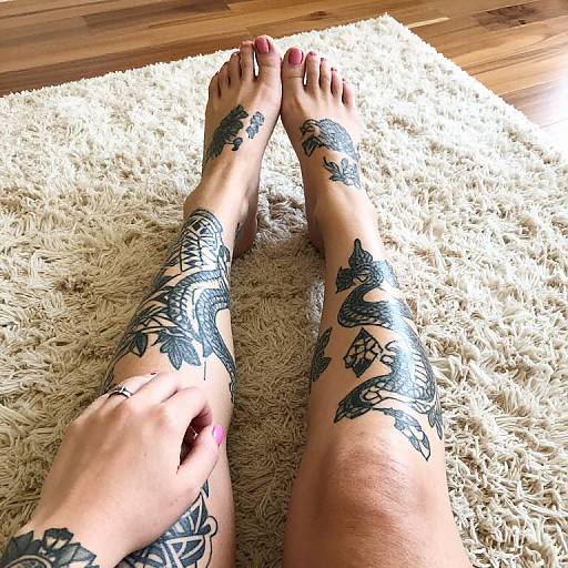 Photograph of tattooed legs with intricate black floral designs on calves and feet, resting on a cream shaggy rug, toes painted red, on