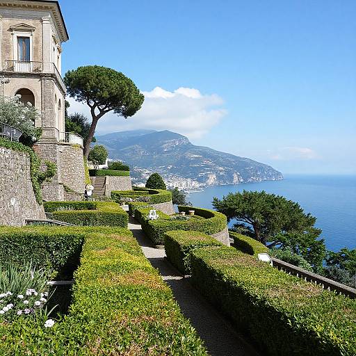 Photograph of a sunlit Italian villa garden with manicured hedges, stone path, tree, and coastal view of blue sea and mountain.