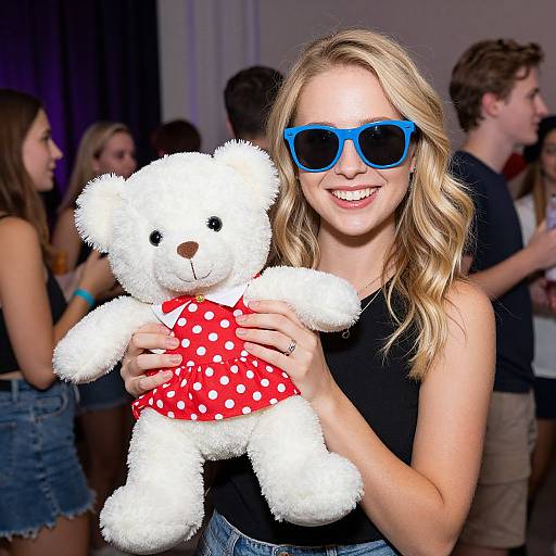 Photograph of a smiling blonde woman with blue sunglasses, holding a white teddy bear in a red polka dot dress, at a social event with