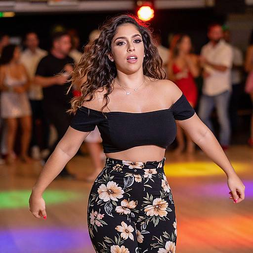 Photograph of a curvy woman with wavy brown hair, wearing an off-shoulder black crop top and floral black skirt, dancing on a