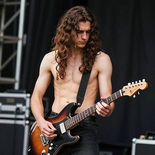 Photograph of a shirtless, muscular man with long brown hair, playing a sunburst electric guitar on stage, wearing black pants and suspenders.