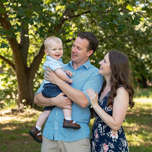 Photograph of a smiling father in a blue checkered shirt, holding a laughing blonde toddler, with a laughing brunette mother in a floral dress, standing