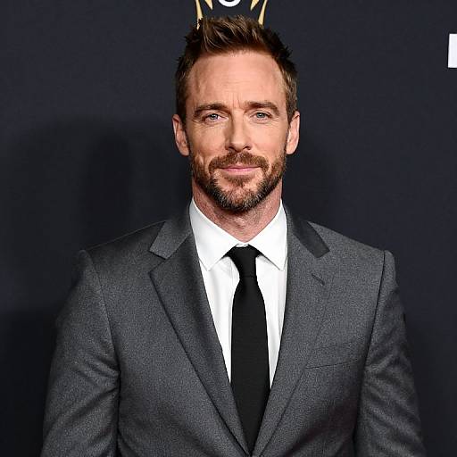 Photograph of a handsome, bearded man with short brown hair, wearing a dark gray suit, white shirt, and black tie, against a black