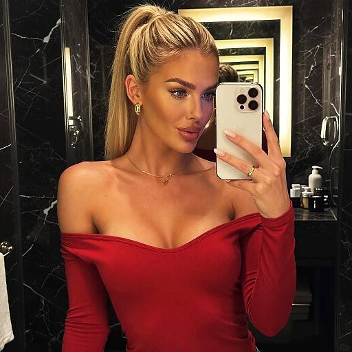 Blonde woman in red off-shoulder dress, gold hoop earrings, taking mirror selfie in black marble bathroom. Photographed with white iPhone.