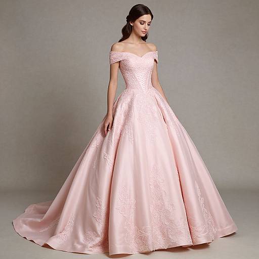 Photograph of a woman in an off-the-shoulder, pink satin ball gown with intricate lace embroidery, standing against a plain gray background.
