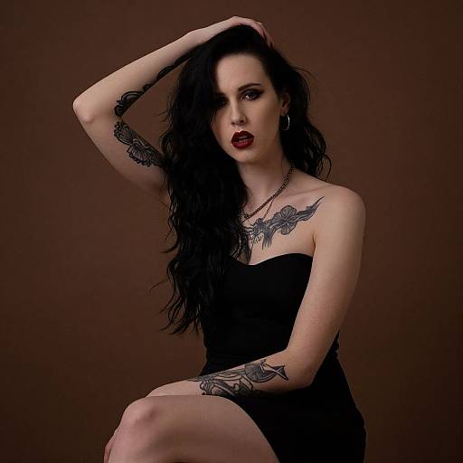 Photograph of a pale-skinned, black-haired woman with dark red lipstick, wearing a black strapless dress, sitting against a brown background. She