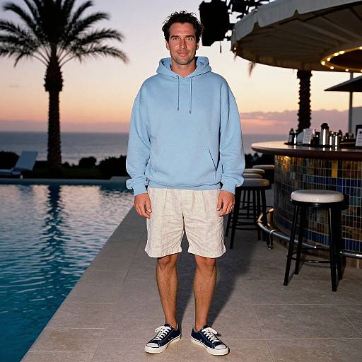Photograph of a man in a light blue hoodie, white shorts, and black sneakers standing by a sunset-lit poolside with palm trees and bar
