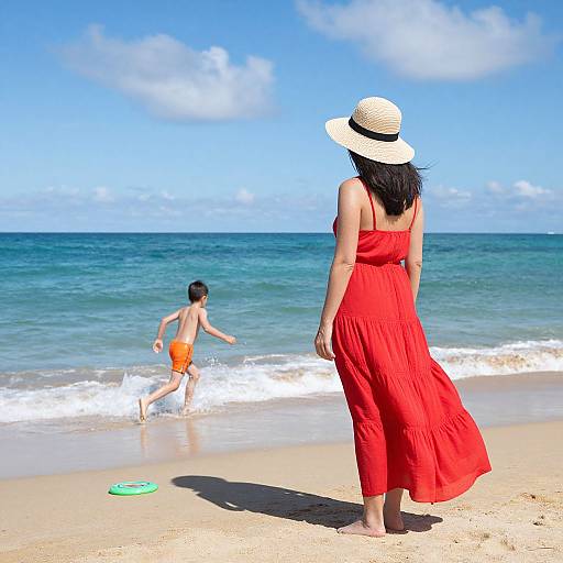 Photograph of a woman in a flowing red dress and sunhat watching a young boy in orange swim trunks play in the ocean waves on a sunny