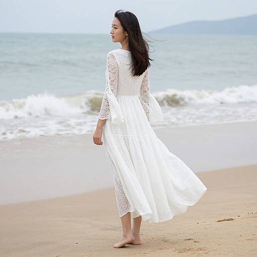 Photograph of a young woman with long black hair, wearing a white lace dress, standing barefoot on a sandy beach with gentle ocean waves in the