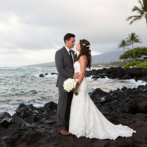 Photograph of a bride in a white lace dress and groom in a gray suit, standing on black rocks by the ocean, holding white flowers, with