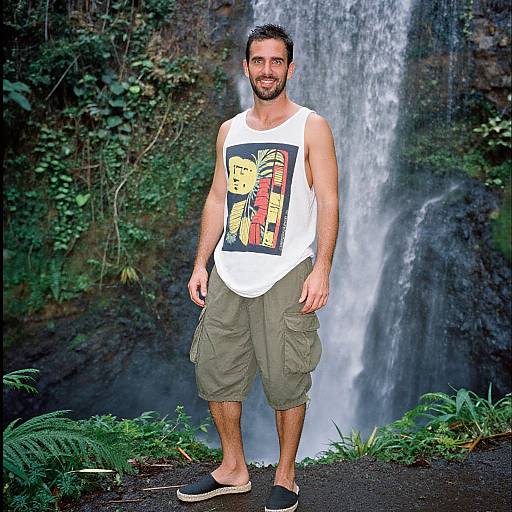 Photograph of a smiling, bearded man with short dark hair, wearing a white tank top with a colorful graphic, olive cargo shorts, and black