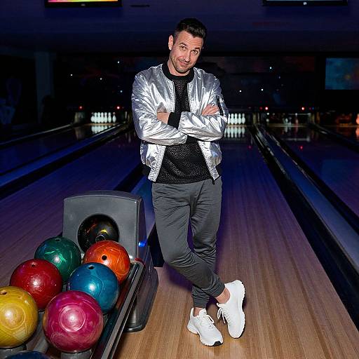 Photograph of a casually dressed man with a short beard, wearing a silver jacket, black shirt, and gray pants, leaning on a bowling ball return