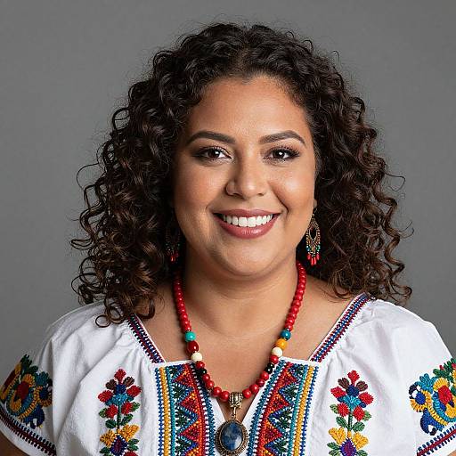 Photograph of a smiling woman with curly dark hair, wearing a white embroidered top, red bead necklace, and matching earrings, against a gray background.