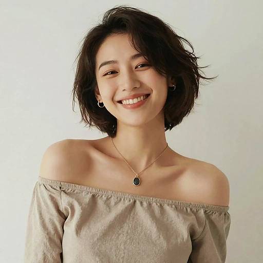 Photograph of a smiling Asian woman with short black hair, wearing an off-shoulder beige top, small hoop earrings, and a pendant necklace,