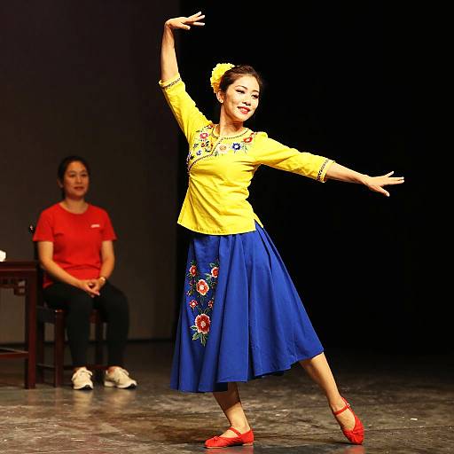 Photograph of a vibrant female dancer in a yellow top and blue skirt, with red shoes and floral embroidery, mid-dance on stage, with a