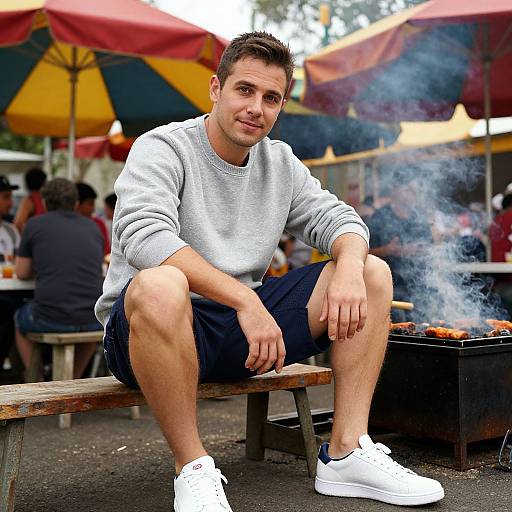 Photograph of a young man with short brown hair, wearing a gray sweater, black shorts, and white sneakers, sitting on a bench, with smoke